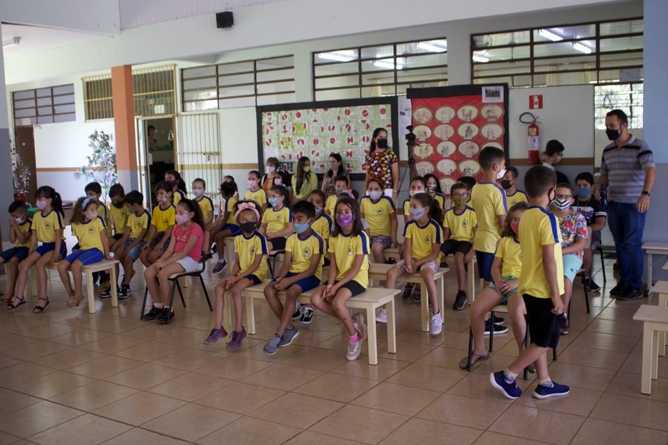 Crianças de uniforme amarelo sentadas em bancos no salão da escola.