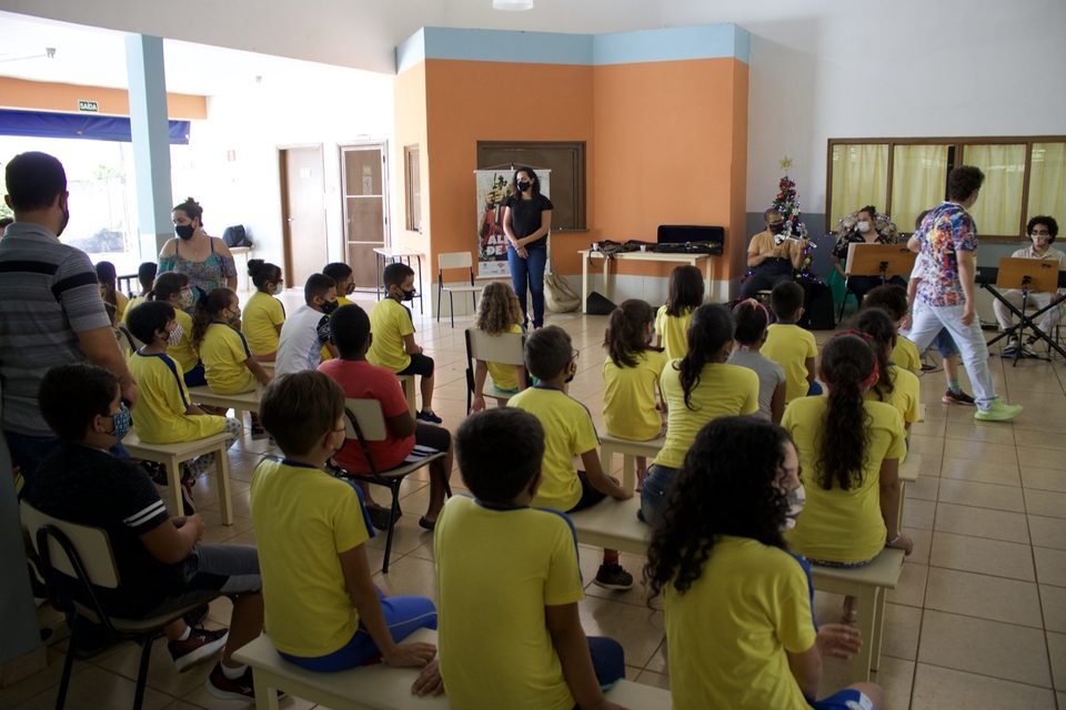 As crianças assistem de perto ao grupo, que toca ao fundo do salão.