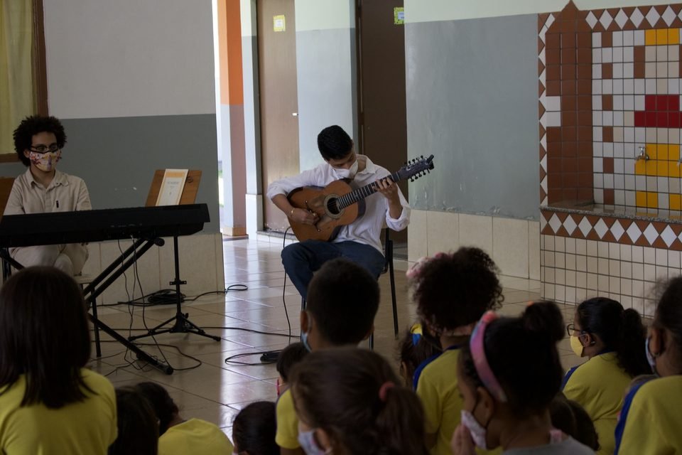 Visto por trás das crianças, um músico toca violão ao lado do teclado.