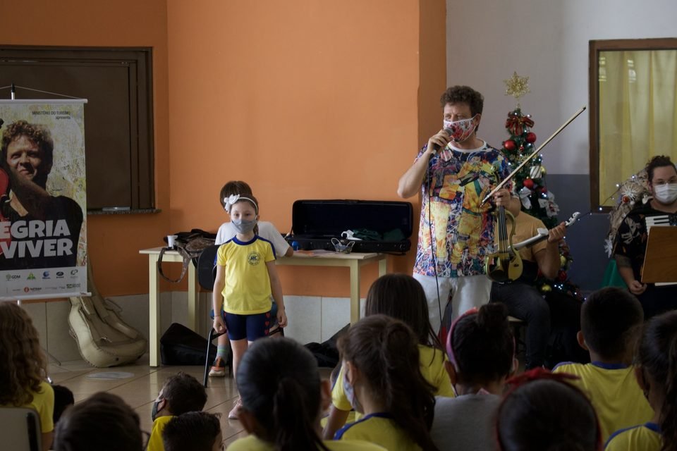Um menino de uniforme amarelo fica de pé ao lado de Roney Marczak, que toca violino.