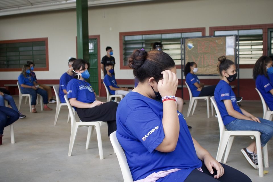 Jovens de camiseta azul e máscara ouvem a apresentação, sentados.