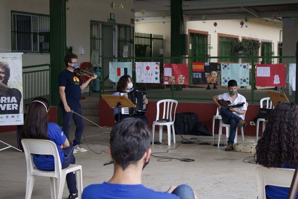 Os músicos tocam diante dos cartazes; jovens assistem em primeiro plano.